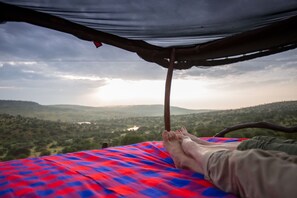 Relax under a rustic shelter with a comfortable bed and scenic views of rolling hills and a distant body of water.