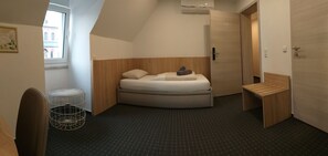 A compact hotel room with a bed, a small wooden bench, a metal basket, and a window with a view of a building. - Hotel Kriemhilde (Worms)