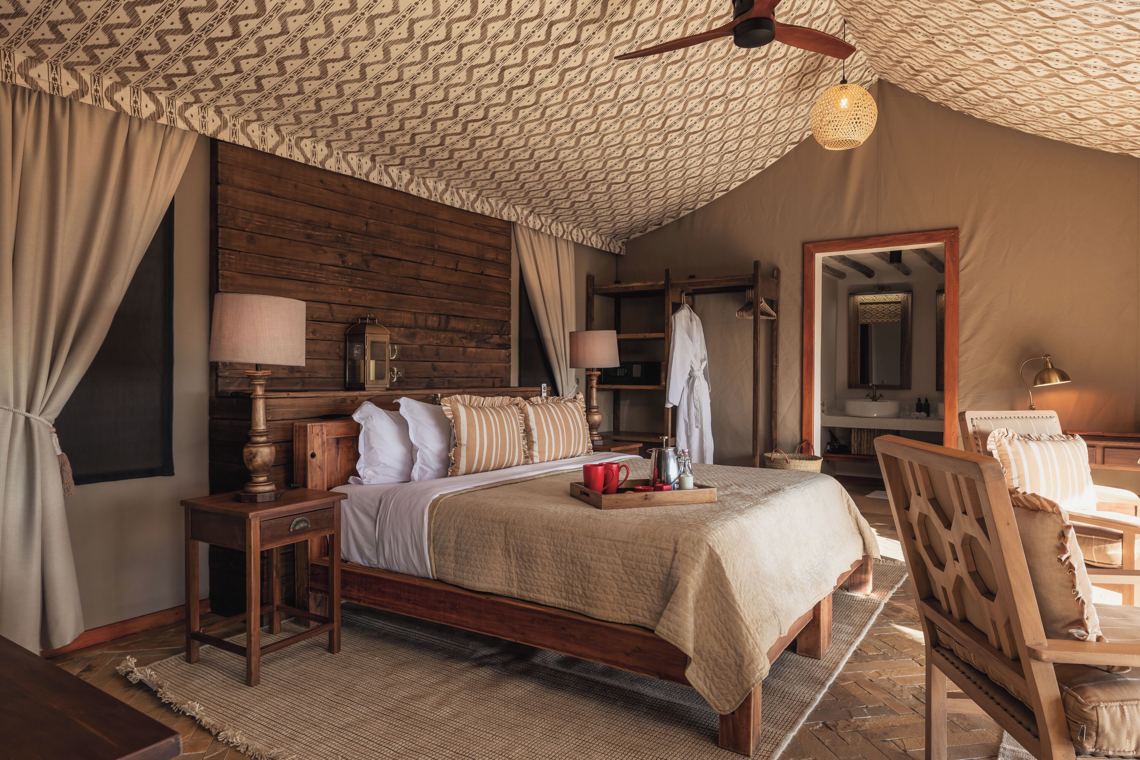 A bedroom with a wooden headboard, a bed with a beige blanket, a bedside table with a lamp, and a mirror on the wall.