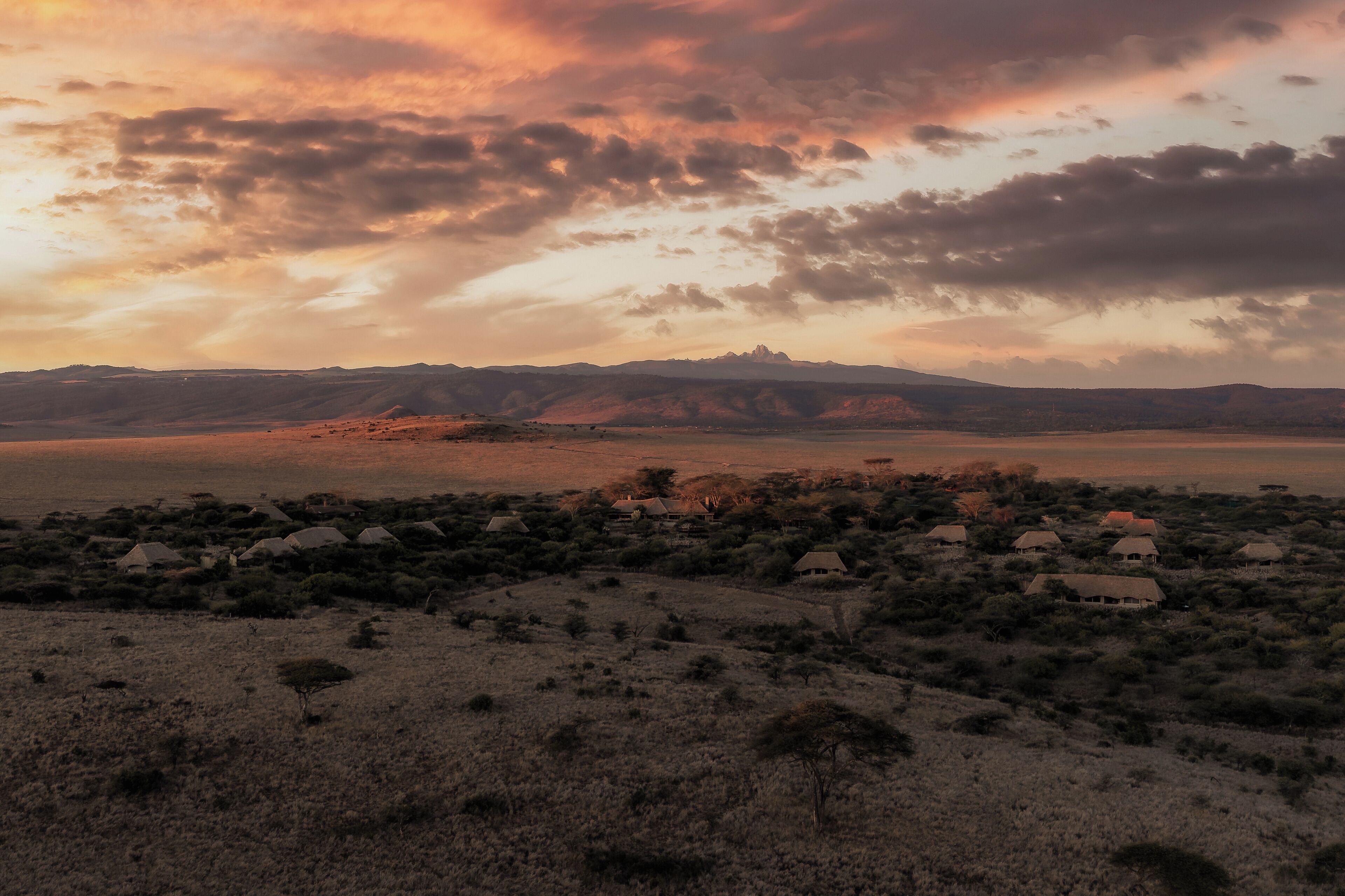 A landscape with a village of thatched-roof houses, a mountain range, and a sunset sky.