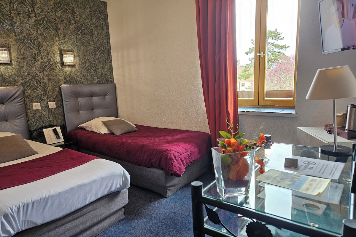 Ein Hotelzimmer mit zwei Betten, einem Glastisch mit Blumenvase, einem Schreibtisch mit Lampe und einem an der Wand montierten Fernseher.