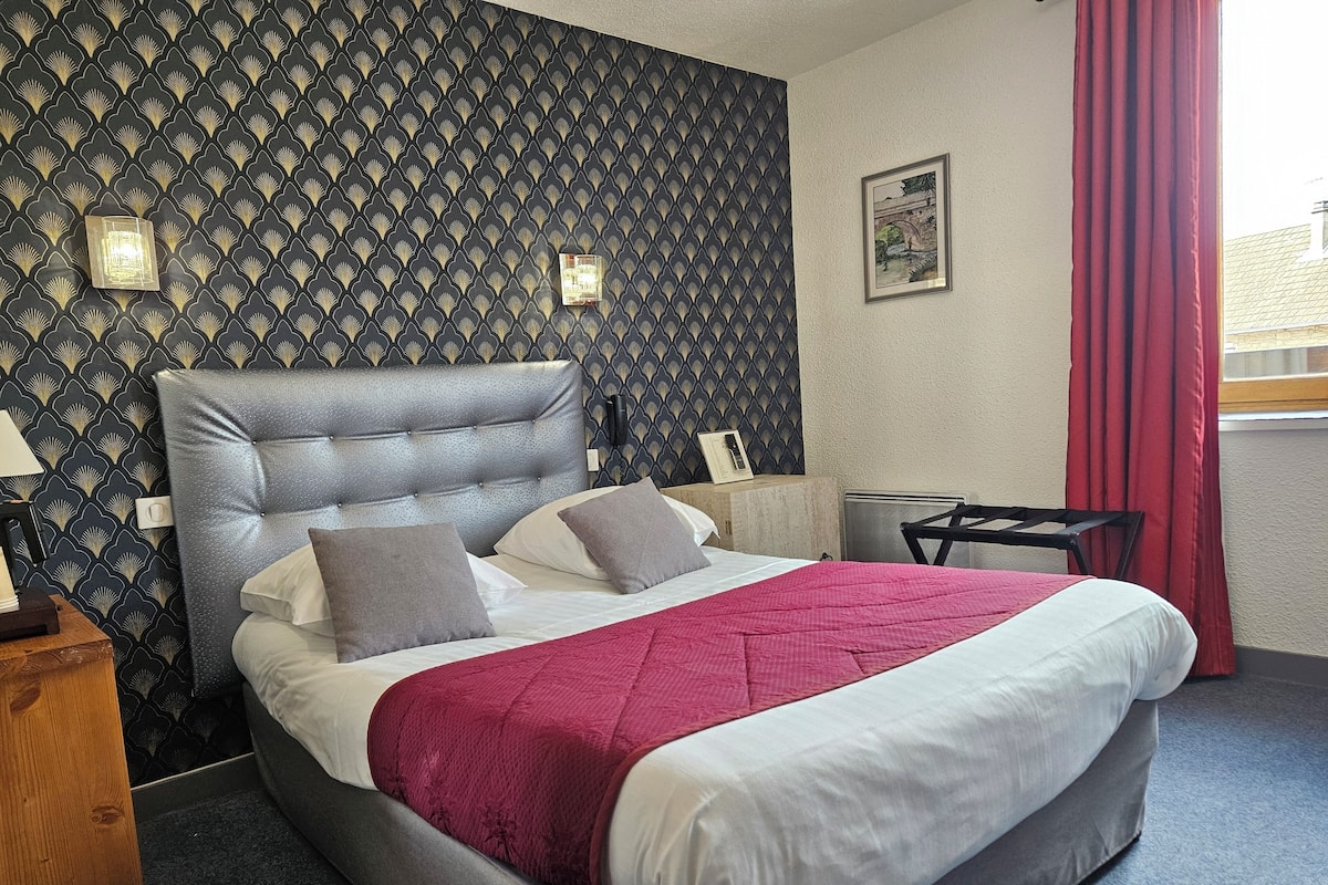 Ein ordentlich bezogenes Bett mit roter Decke, einem grauen gepolsterten Kopfteil und einer Wand mit geometrischem Muster.