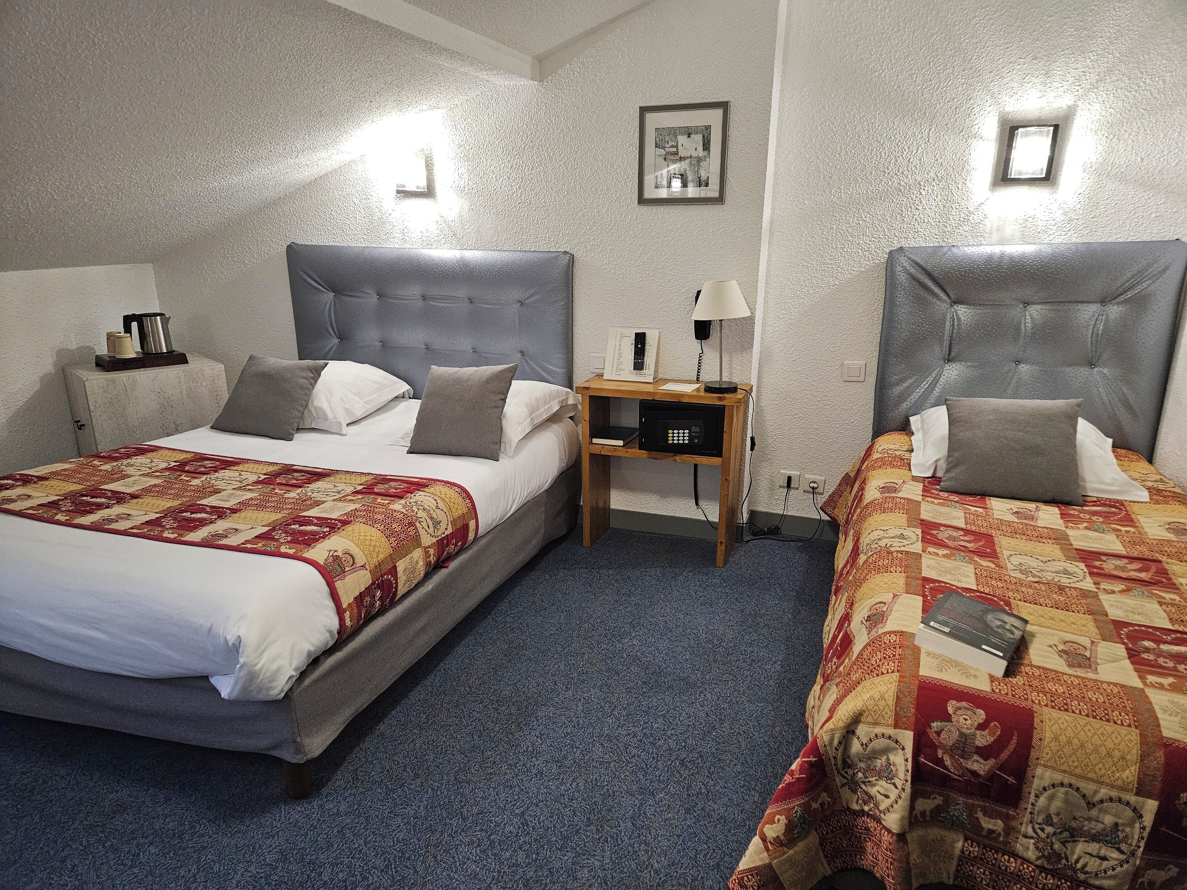A hotel room with two beds, each with a grey headboard, a wooden nightstand, and a lamp. There is a quilt on each bed, and a framed picture on the wall.