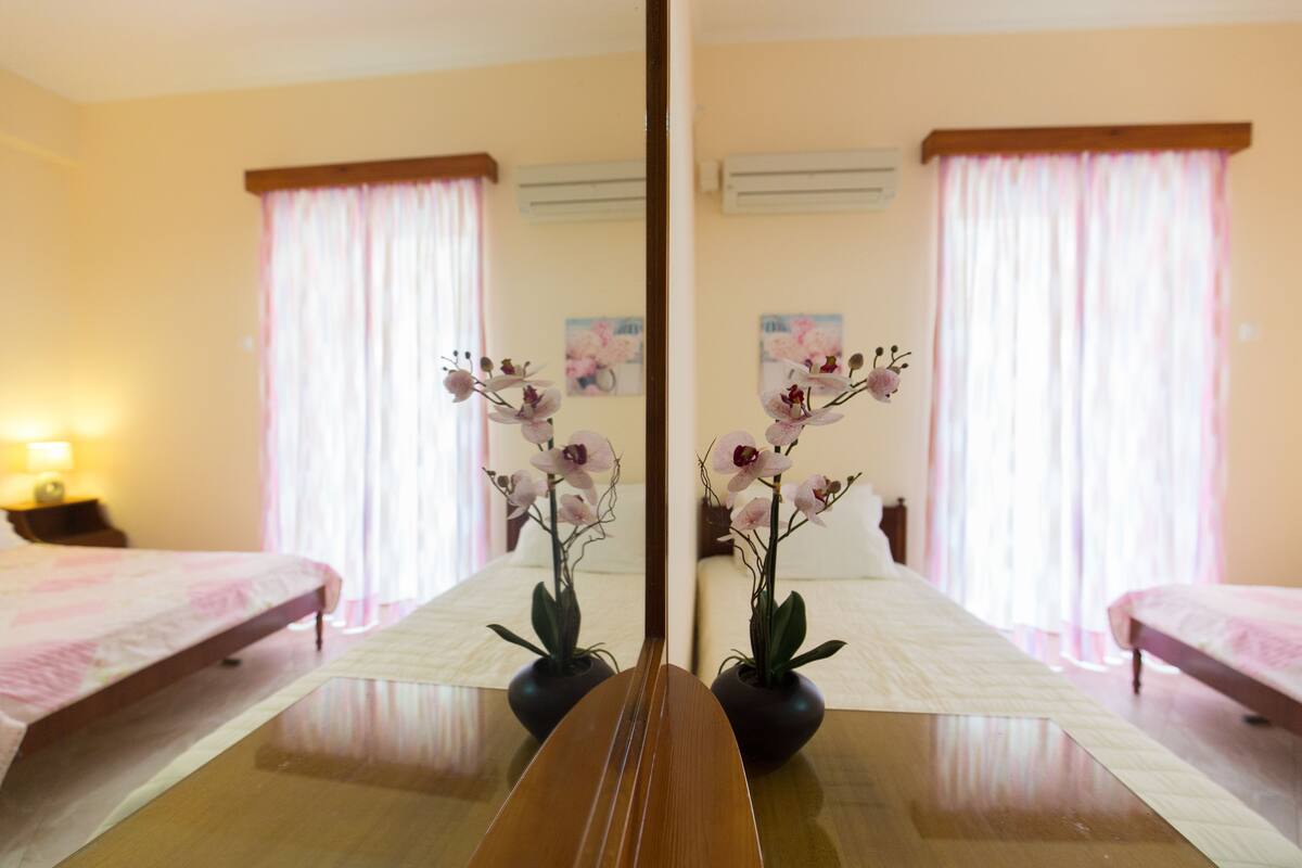 A hotel room with a bed, a table with a vase of flowers, and a mirror reflecting the room.