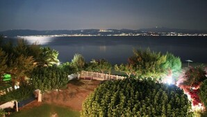 A nighttime view of a coastal area with a calm sea, a distant cityscape, and a well-lit garden with trees and a fence. - Pansion Akrogiali (Eretria)