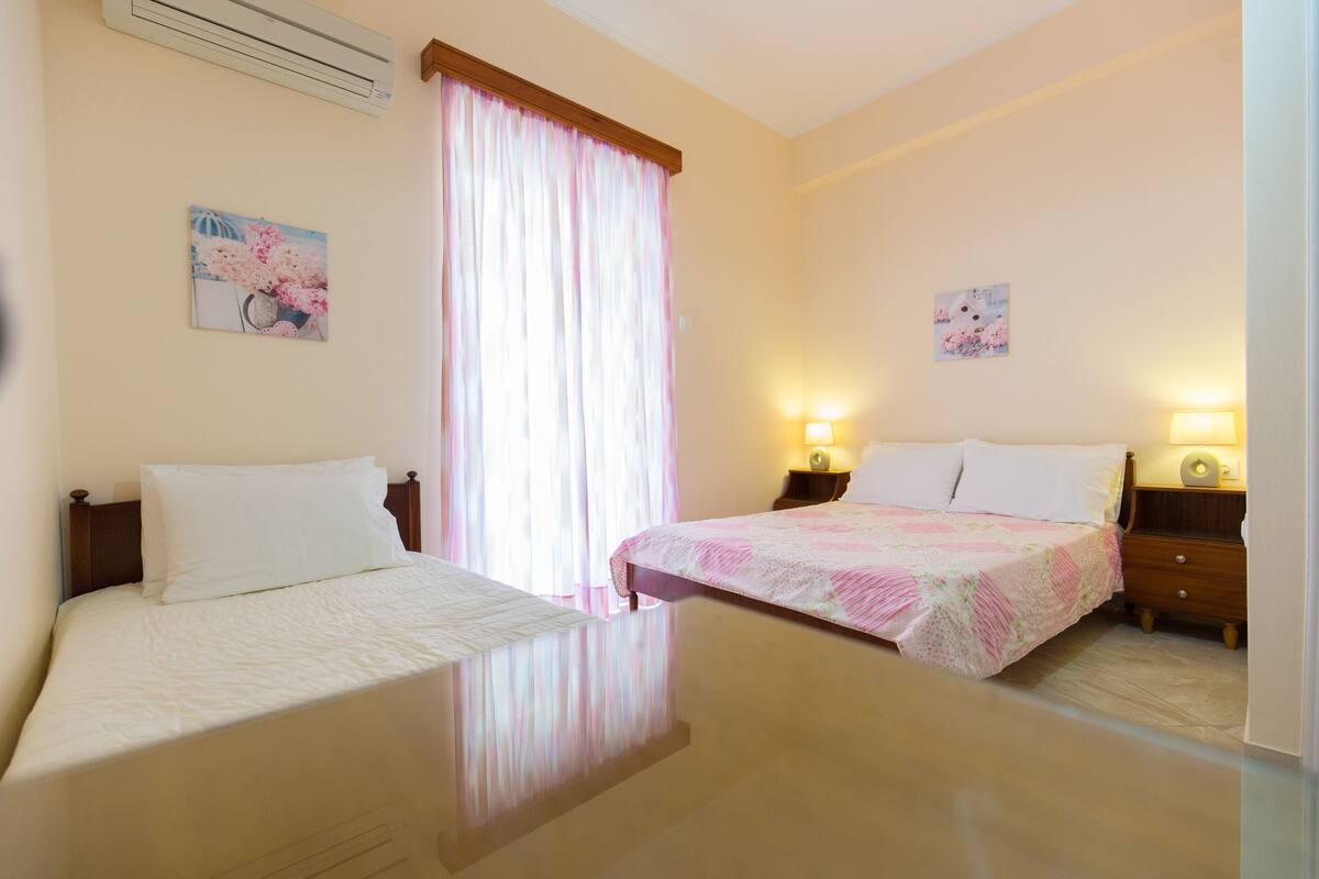 A hotel room with two beds, a large window with curtains, wall-mounted air conditioning, bedside lamps, and artwork on the walls.