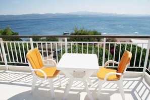 Ein Balkon mit zwei weißen Stühlen und einem kleinen Tisch, der Blick auf die Küste mit einem Schiff in der Ferne gewährt.