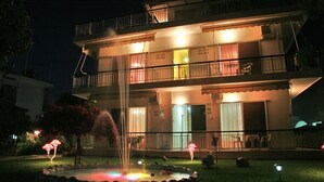 A multi-story hotel with a balcony on the second floor, a well-lit entrance, and a small outdoor fountain. - Pansion Akrogiali (Eretria)