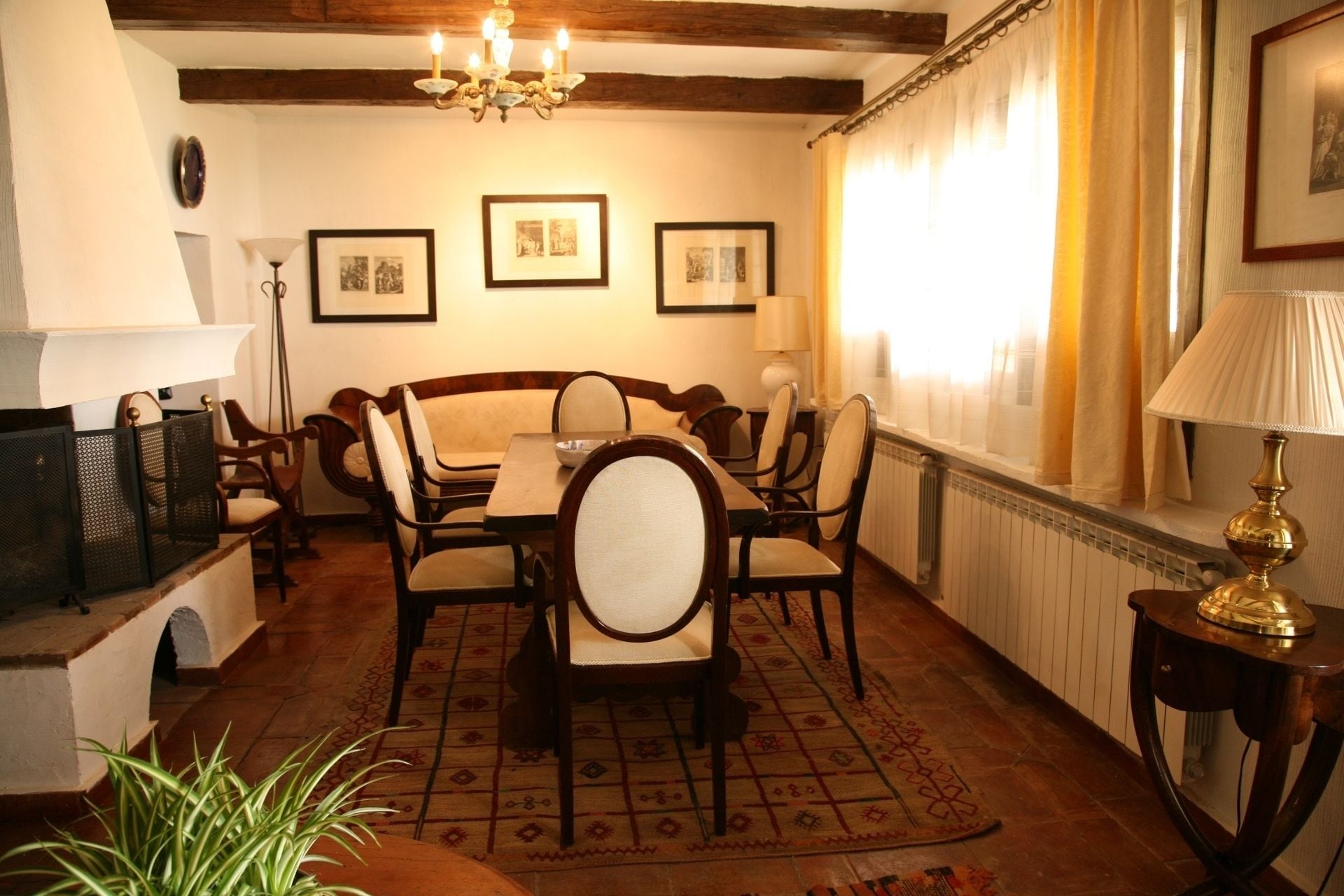 A dining room with a wooden table and chairs, framed pictures on the wall, a chandelier, and a window with curtains.