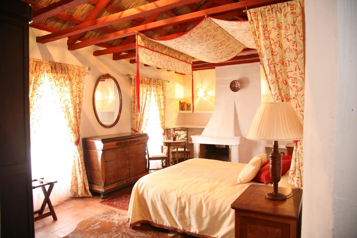 Un dormitorio acogedor con una cama de madera, un tocador, una silla y una mesita de noche. Hay una ventana con cortinas, un espejo y una lámpara en la mesita de noche.
