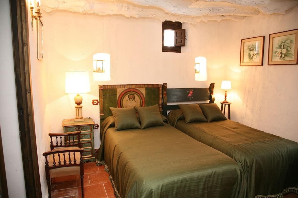 Una habitación con dos camas, una silla de madera y una pequeña mesa con una lámpara.