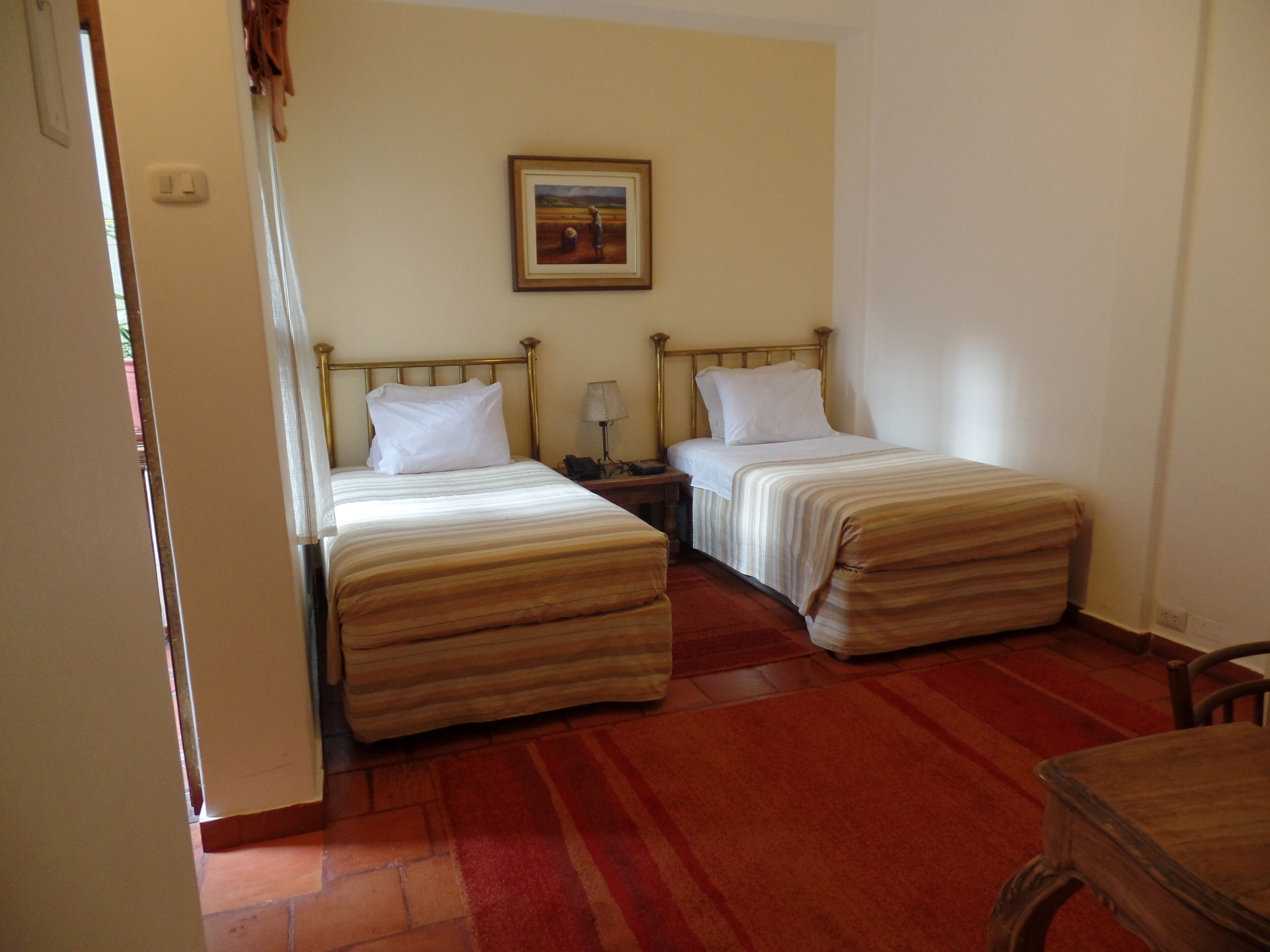 Quarto com duas camas de solteiro, uma mesa de cabeceira com abajur, um quadro na parede e uma cadeira de madeira.
