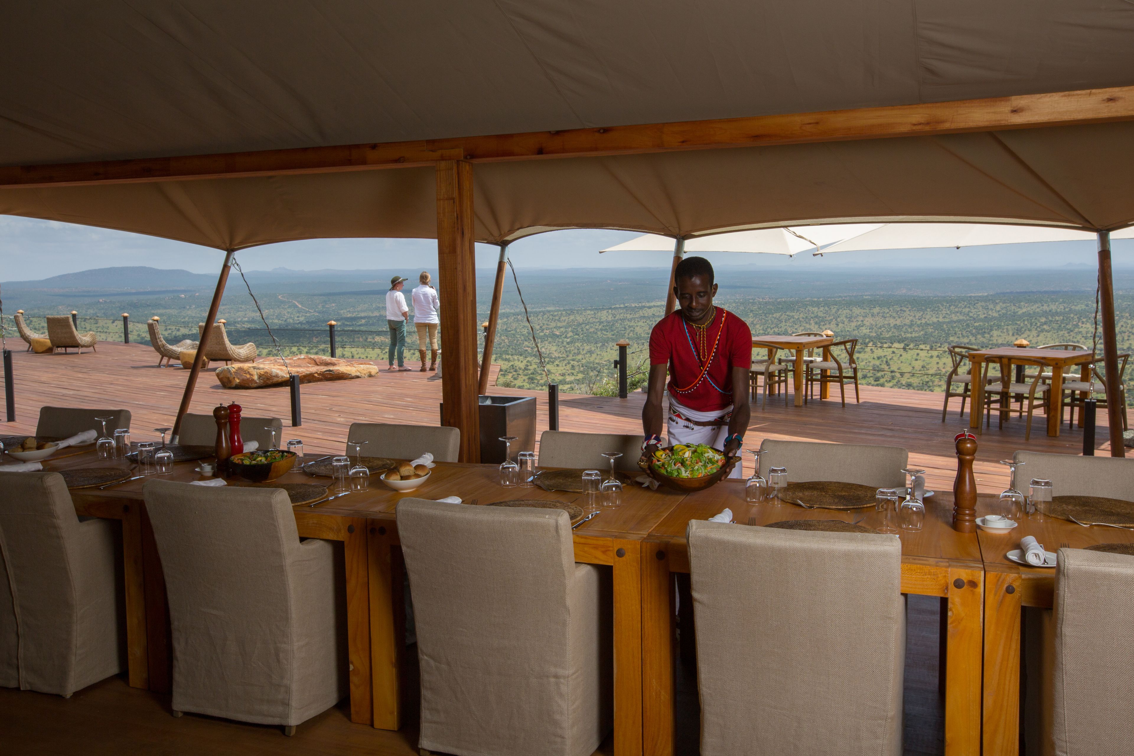 Un espacio de comedor al aire libre con mesas y sillas, una persona sirviendo comida, y una vista panorámica de fondo.