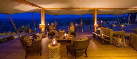 A tented outdoor dining area with wooden furniture, tables, and chairs, and a view of the landscape.