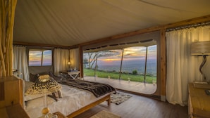 A bedroom with a large bed, a wooden headboard, a bedside table with a lamp, and a view of the ocean at sunset.