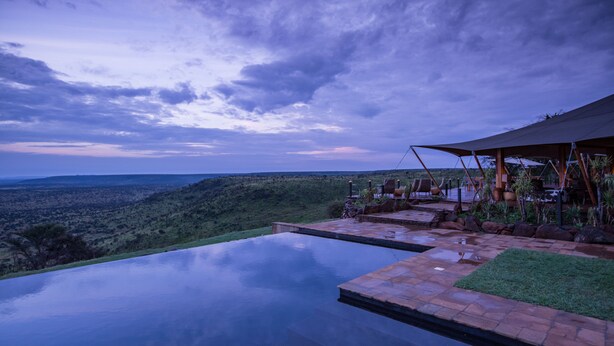 An infinity pool with a view of a hilly landscape and a covered outdoor seating area with chairs and tables.