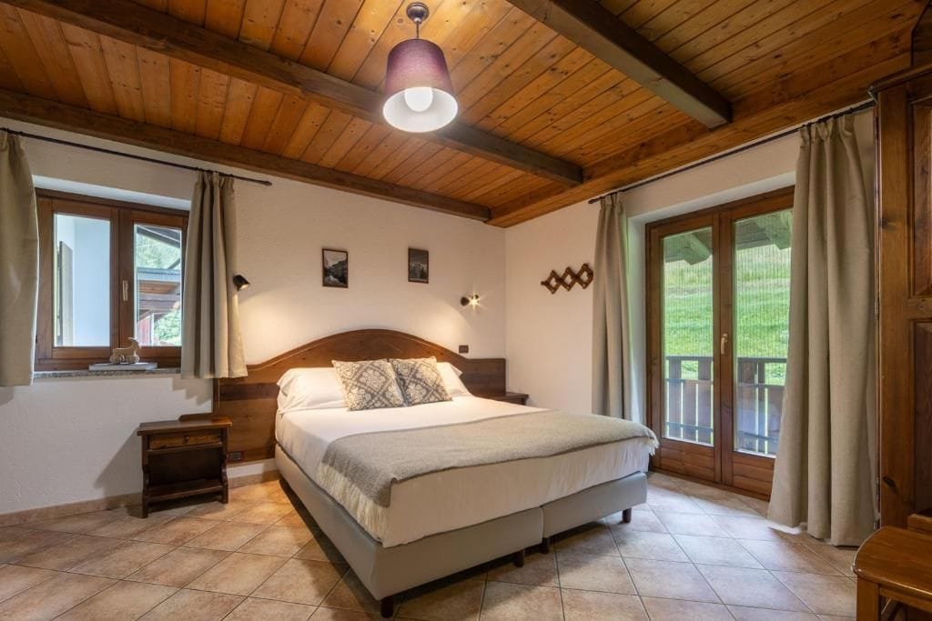 A bedroom with a wooden ceiling, a bed with a headboard, a nightstand, a window with curtains, and a view of greenery outside.