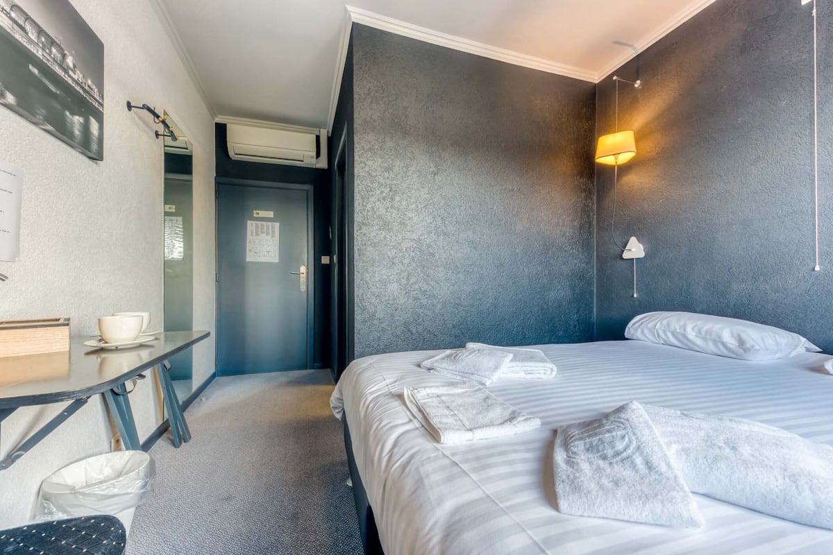 Habitación de hotel con cama, escritorio, silla, cesto de basura y lámpara de pared.