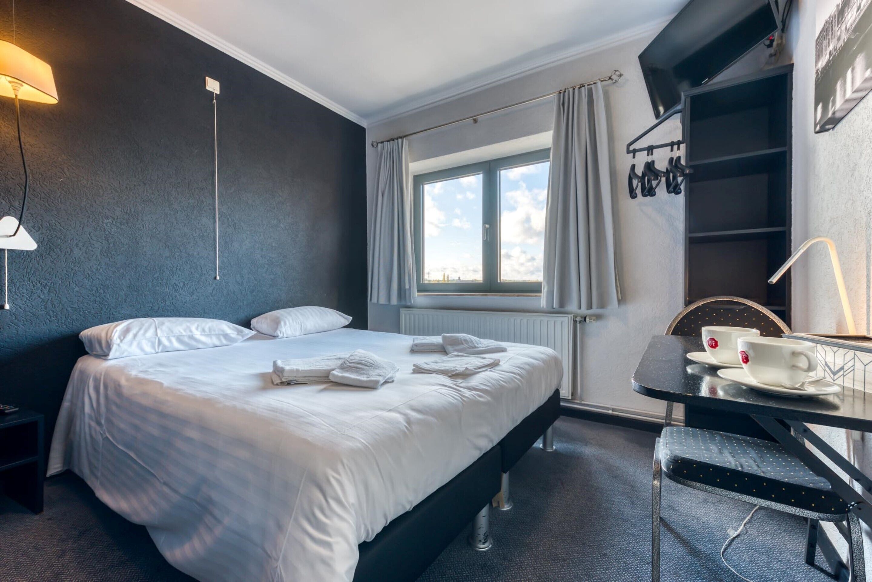 Una habitación de hotel con cama, escritorio, silla, ventana y televisor en la pared.