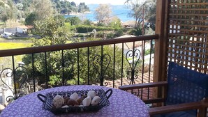 A balcony with a table covered by a purple floral tablecloth, a basket of fruits, and a view of a lake and houses in the background.