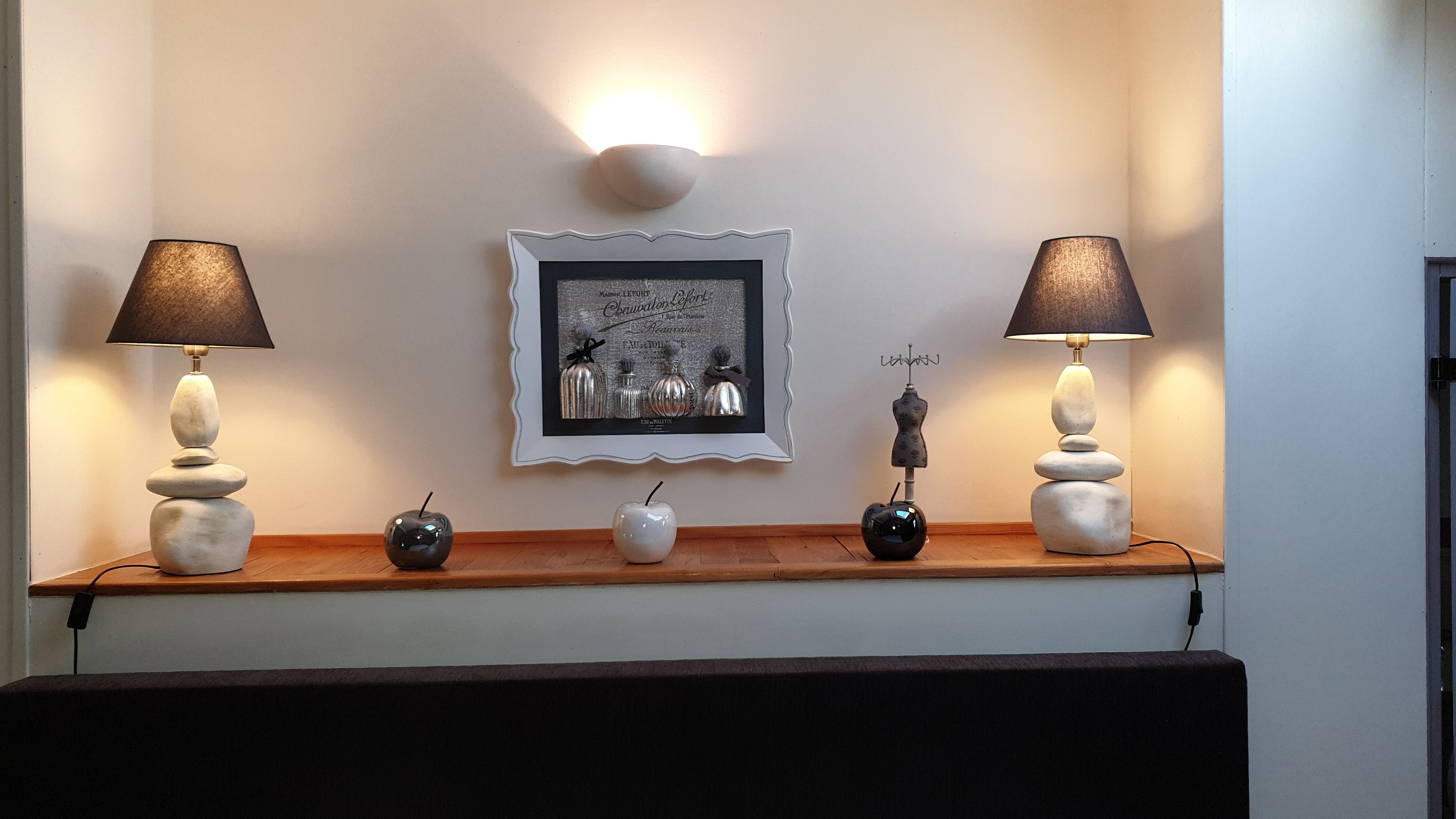 Une pièce avec une étagère en bois où sont exposés des objets décoratifs, notamment deux lampes avec des abat-jour noirs, un tableau encadré et un petit vase.
