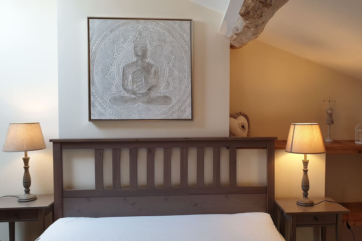 Un dormitorio con cabecera de madera, una obra de Buda enmarcada, dos lámparas de noche, una mesita de noche con lámpara, una estantería con objetos decorativos y un techo de madera.