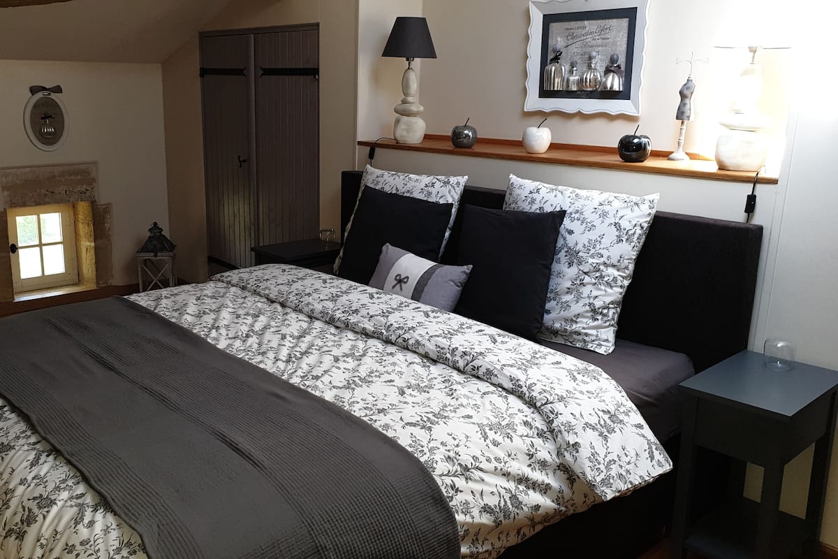 Un dormitorio con una cama grande, cabecera de madera, mesita de noche, lámpara, un cuadro enmarcado y una ventana con vistas al exterior.