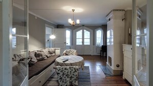 A room with a wooden floor, a sofa with patterned cushions, a round table with a chair, and a chandelier. - Klubbensborg (Hägersten)