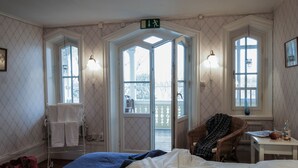 A hotel room with a bed, a chair, a table with a bottle and glass, a towel rack, and a door with a sign. - Klubbensborg (Hägersten)