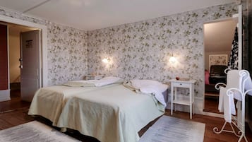 Un dormitorio con papel tapiz floral, una cama con dos almohadas, mesitas de noche, una silla blanca y un piano al fondo.