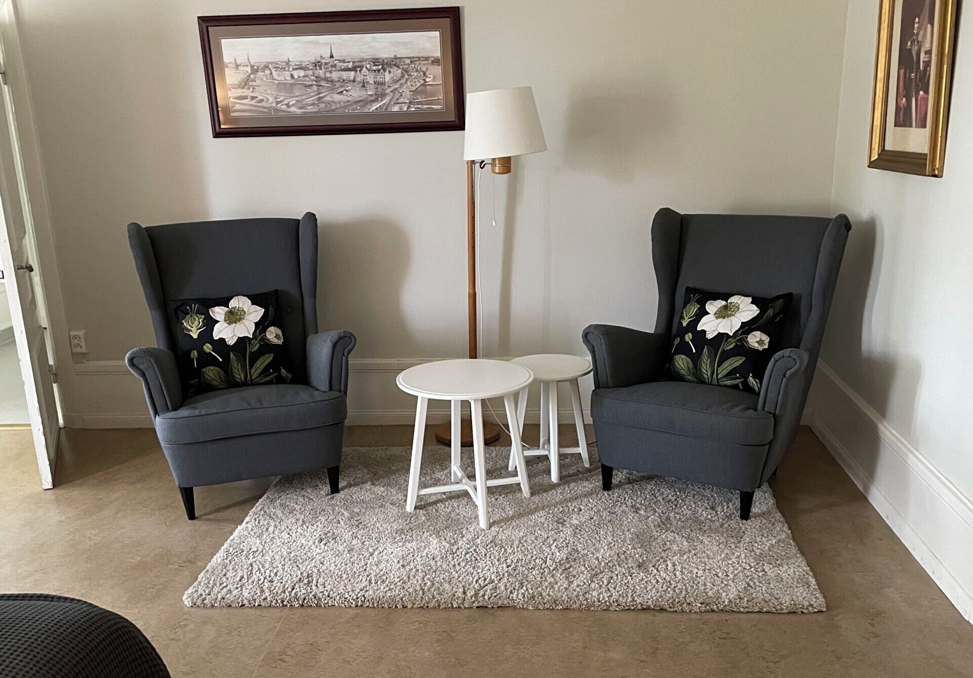 A room with two grey armchairs with floral cushions, a white round table, a tall floor lamp, and a framed cityscape picture on the wall.