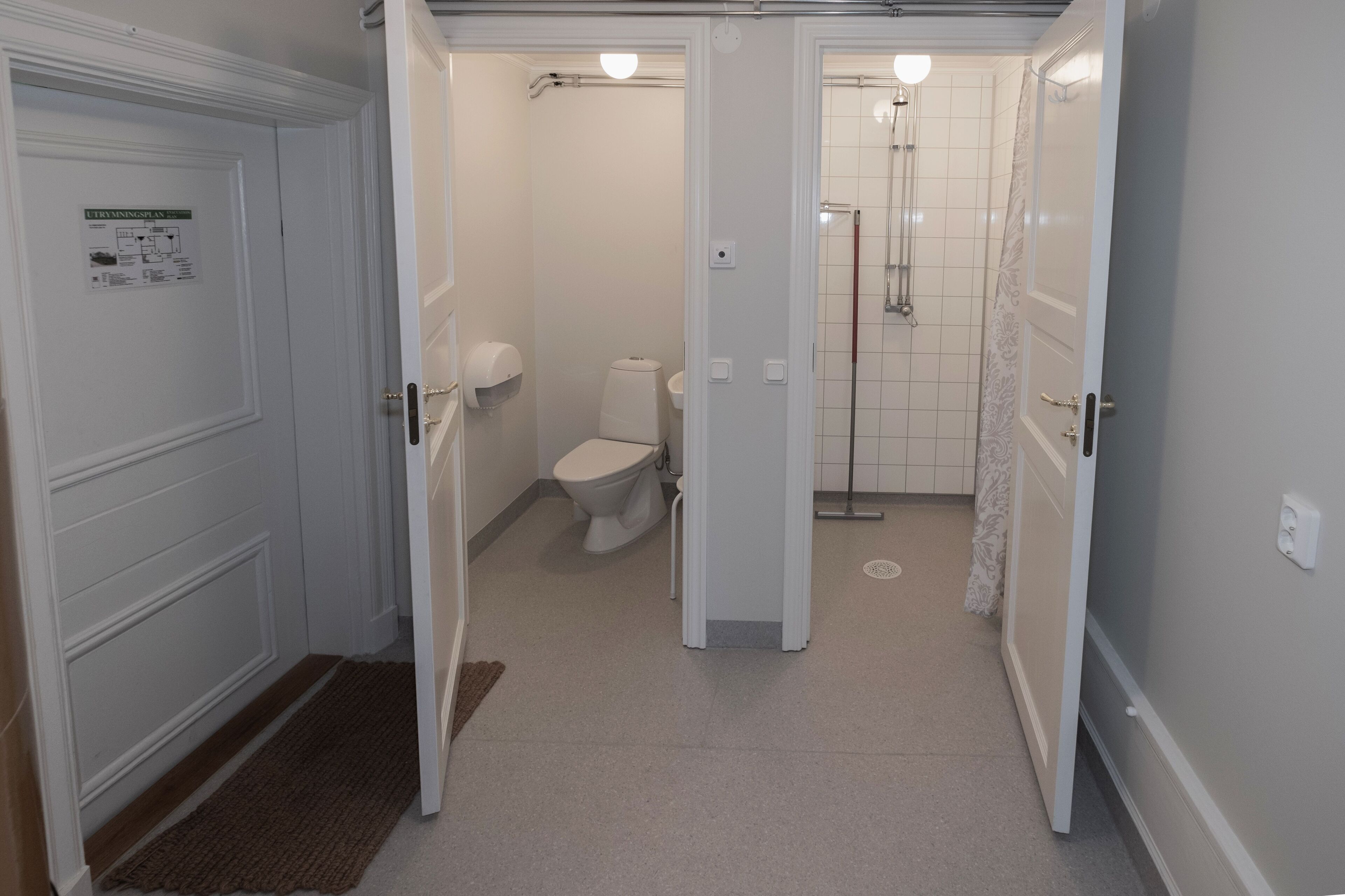 Corredor estreito que dá para um banheiro com vaso sanitário, chuveiro e uma porta com uma placa.