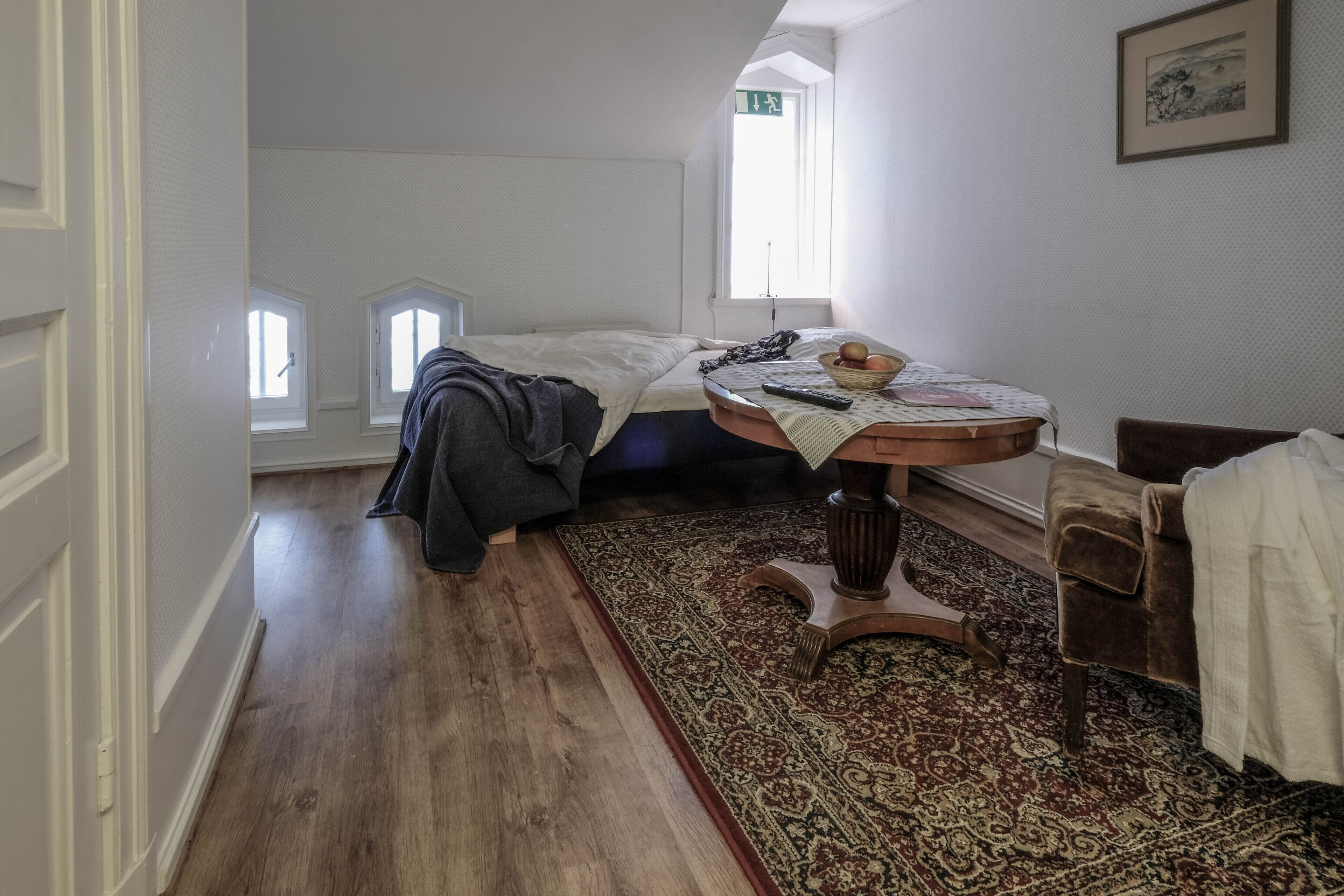 A bedroom with a bed, a small table with a basket and remote, a chair, and a window with a view.