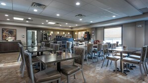 A spacious dining area with multiple tables and chairs, a kitchen area with a refrigerator and counter, and a large window providing natural light. - Timber Ridge Inn and Suites (Fox Creek)