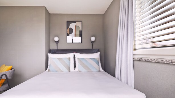 A neatly made bed with striped pillows, a wall-mounted abstract art piece, a lamp, and a window with blinds. - Numa Zurich Turi (Zürich)