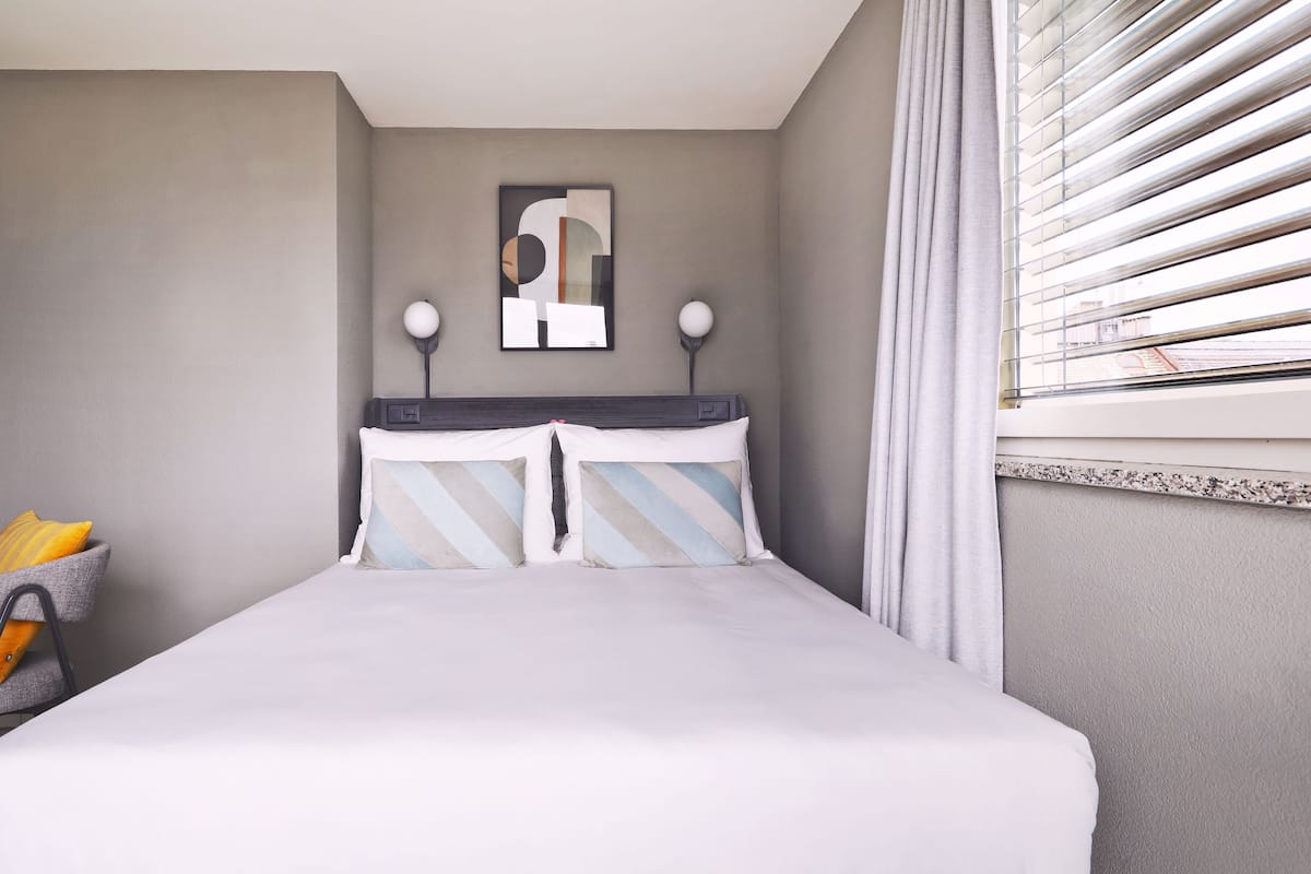 A neatly made bed with striped pillows, a wall-mounted abstract art piece, a lamp, and a window with blinds.