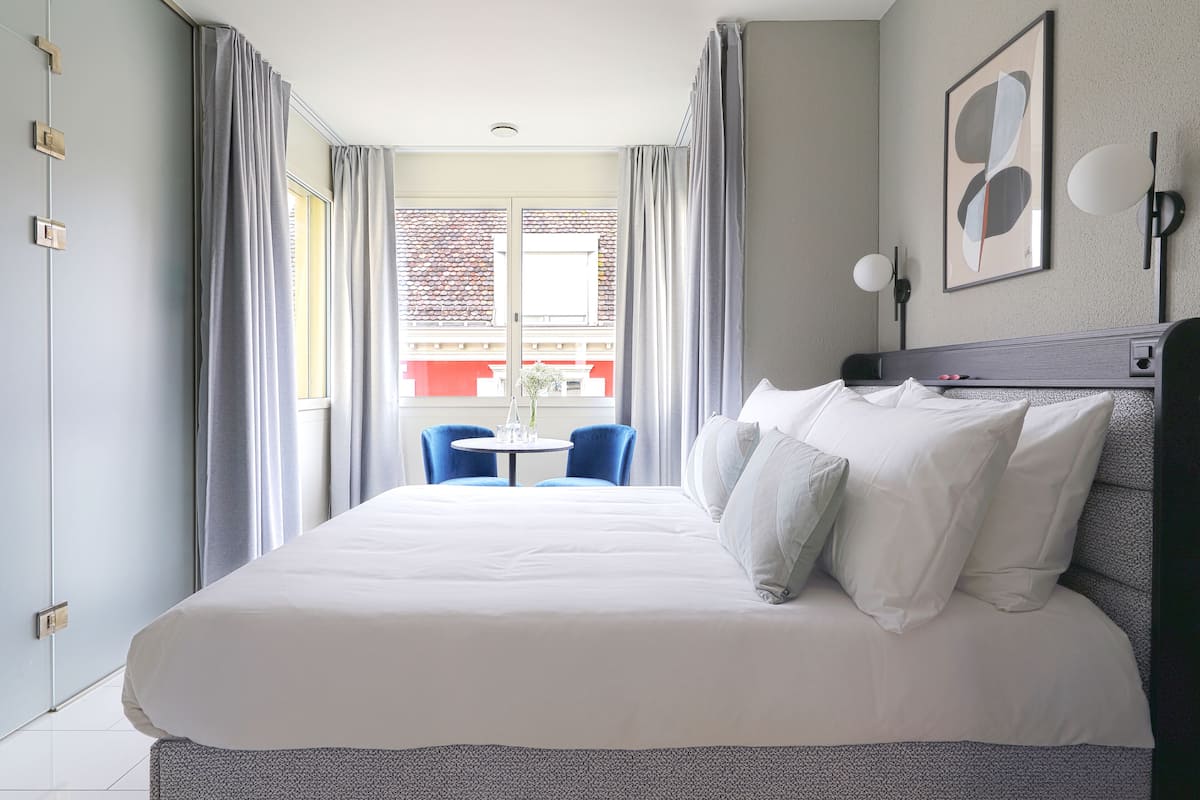 A hotel room with a large bed, a small table, blue chairs, and a window with curtains.