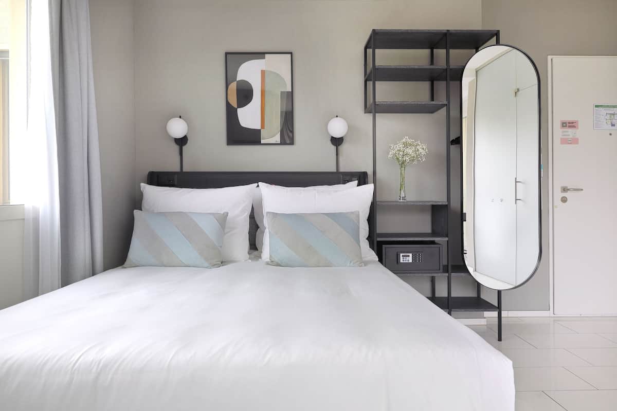 A neatly made bed with white and striped pillows, a bedside table with a lamp, a wall-mounted abstract painting, and a wardrobe with a mirror.