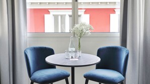 A room with a round table and two blue chairs, a vase with flowers, and a window with a view of a building with red and white colors. - Numa Zurich Turi (Zürich)
