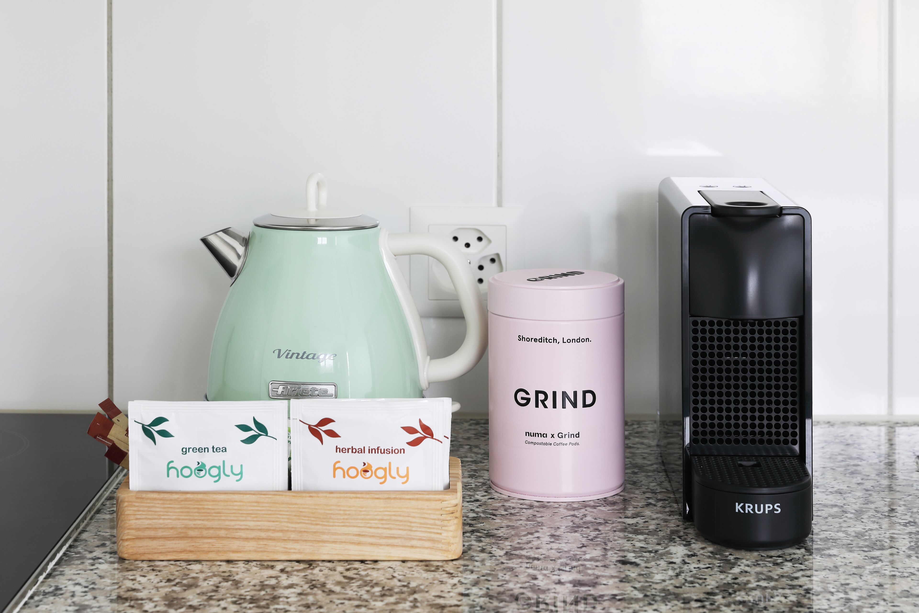 Uma bancada de cozinha com uma chaleira verde, um recipiente rosa para cápsulas de café, um moedor de café preto e duas caixas com a etiqueta 'hoogly'.