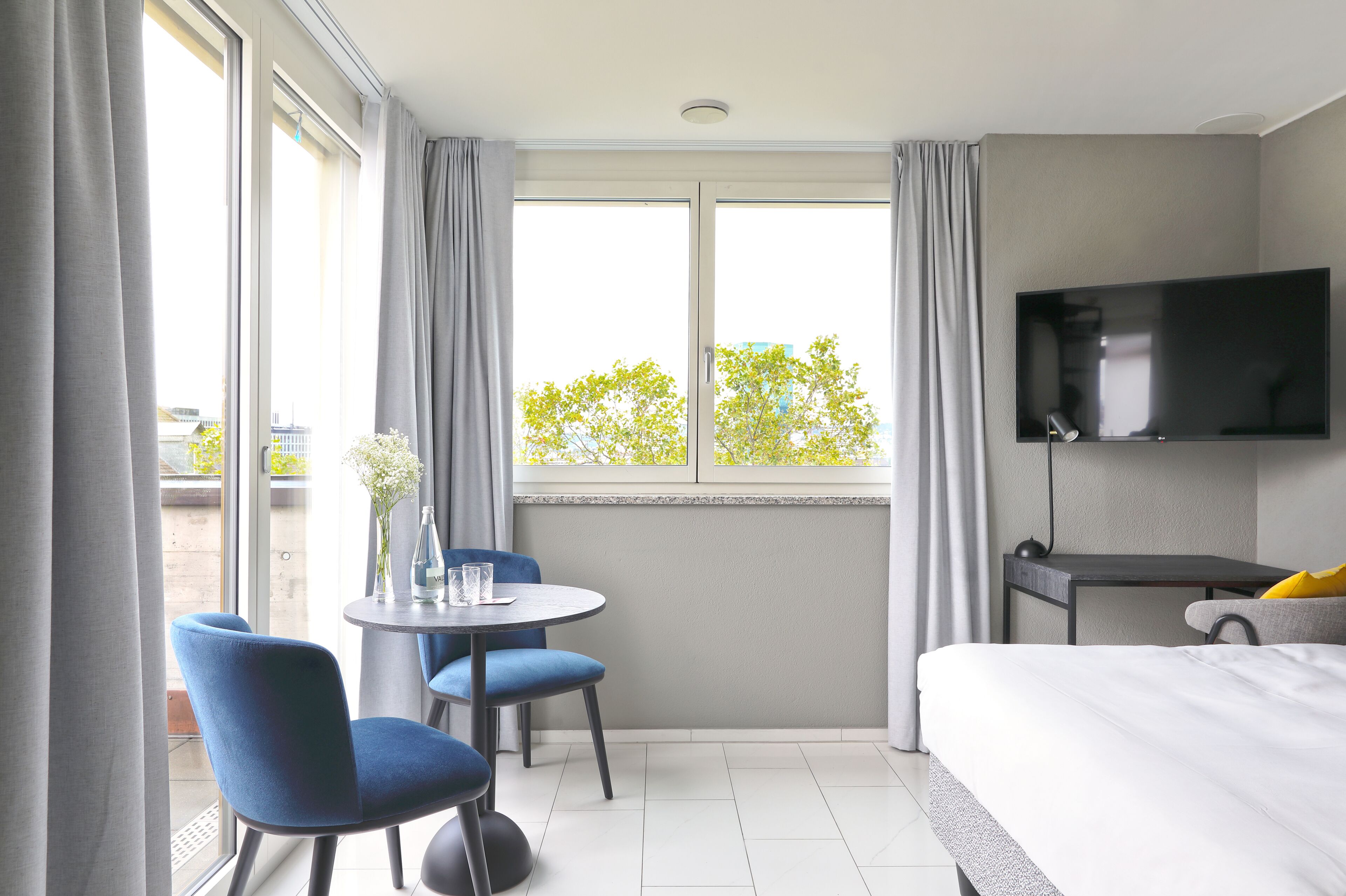 A hotel room with a bed, a TV mounted on the wall, a small table with chairs, and a window with curtains.