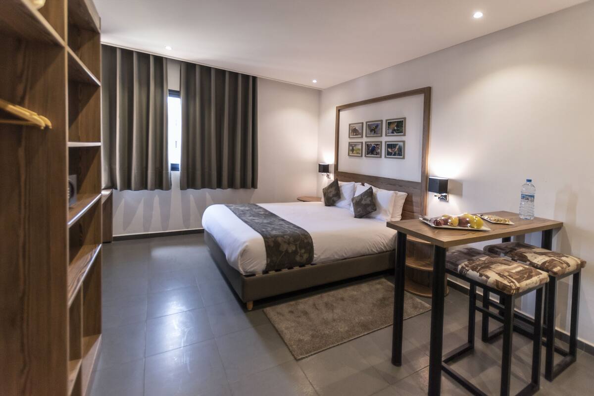 A modern hotel room with a large bed, a wooden side table, a small table with a water bottle, and a bench.