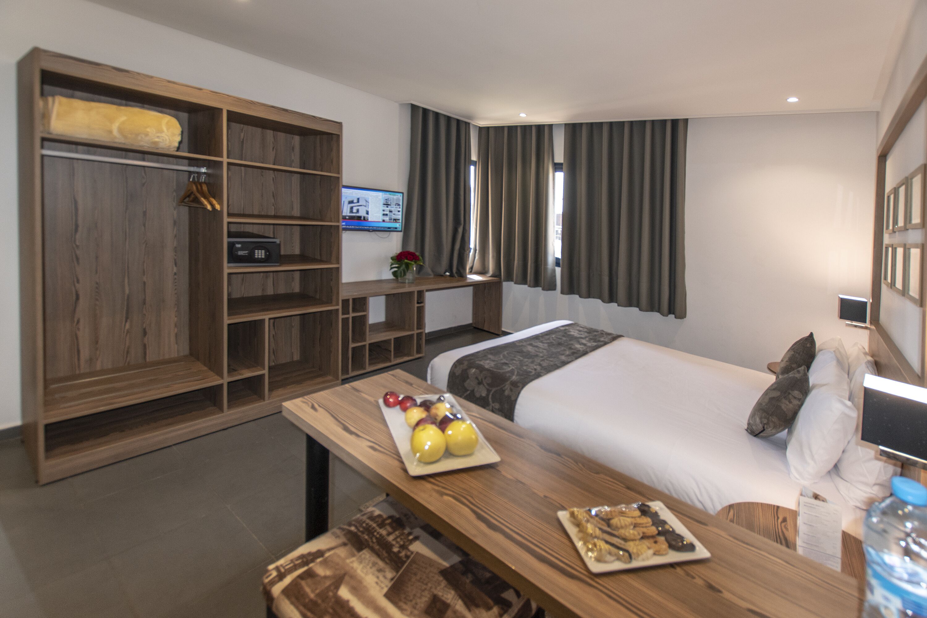Une chambre d’hôtel moderne avec un grand lit, une armoire en bois, un petit bureau avec un plateau de fruits et un téléviseur fixé au mur.