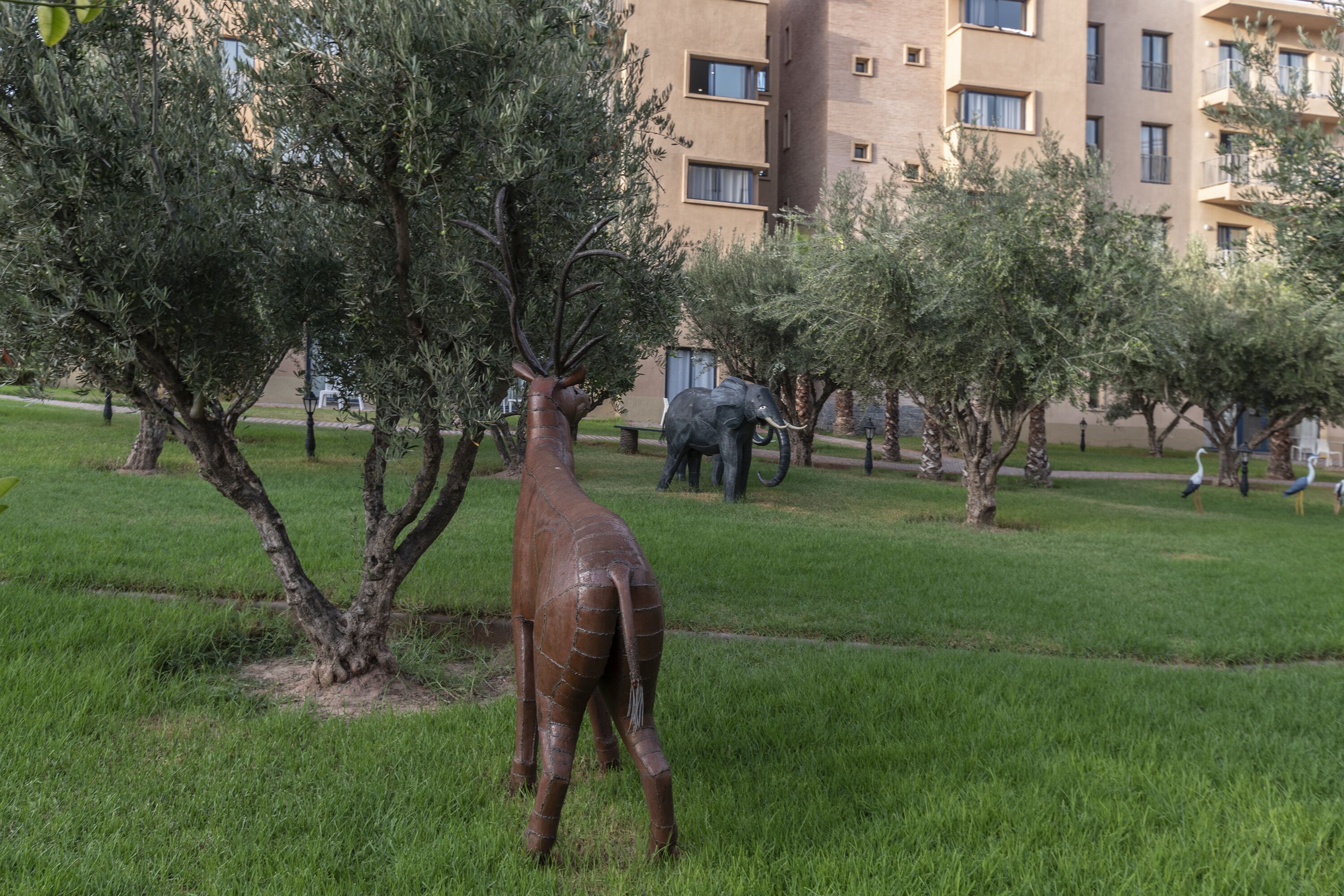 A sculpture of a giraffe in a grassy area with trees and a building in the background.