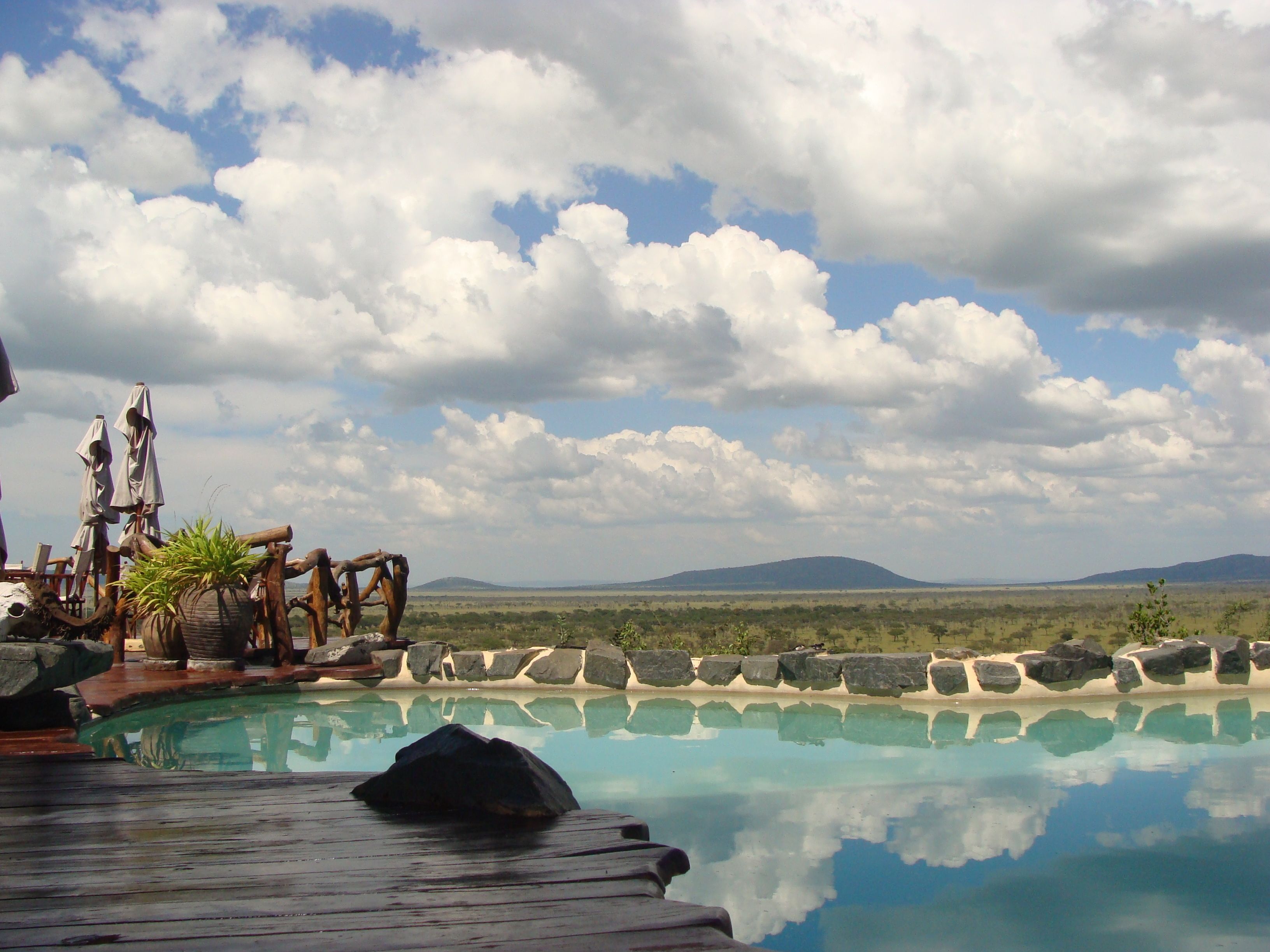 Une piscine dotée d’une terrasse en bois, entourée de rochers et de plantes, offrant une vue sur un paysage lointain et des montagnes sous un ciel nuageux.