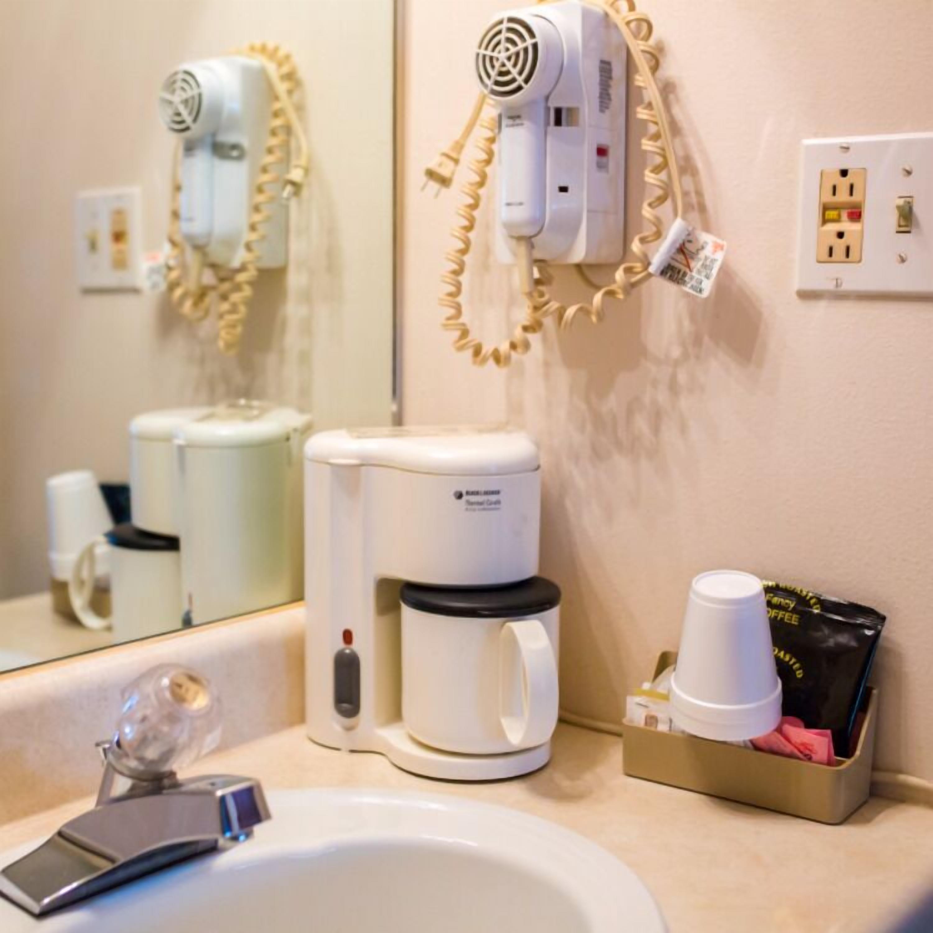 A hotel bathroom with a sink, a coffee machine, a hairdryer, and a cup holder with coffee cups.
