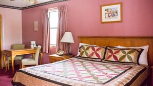 Habitación de hotel con cama, escritorio, silla, mesita de noche, lámpara, ventilador de techo y un cuadro en la pared.