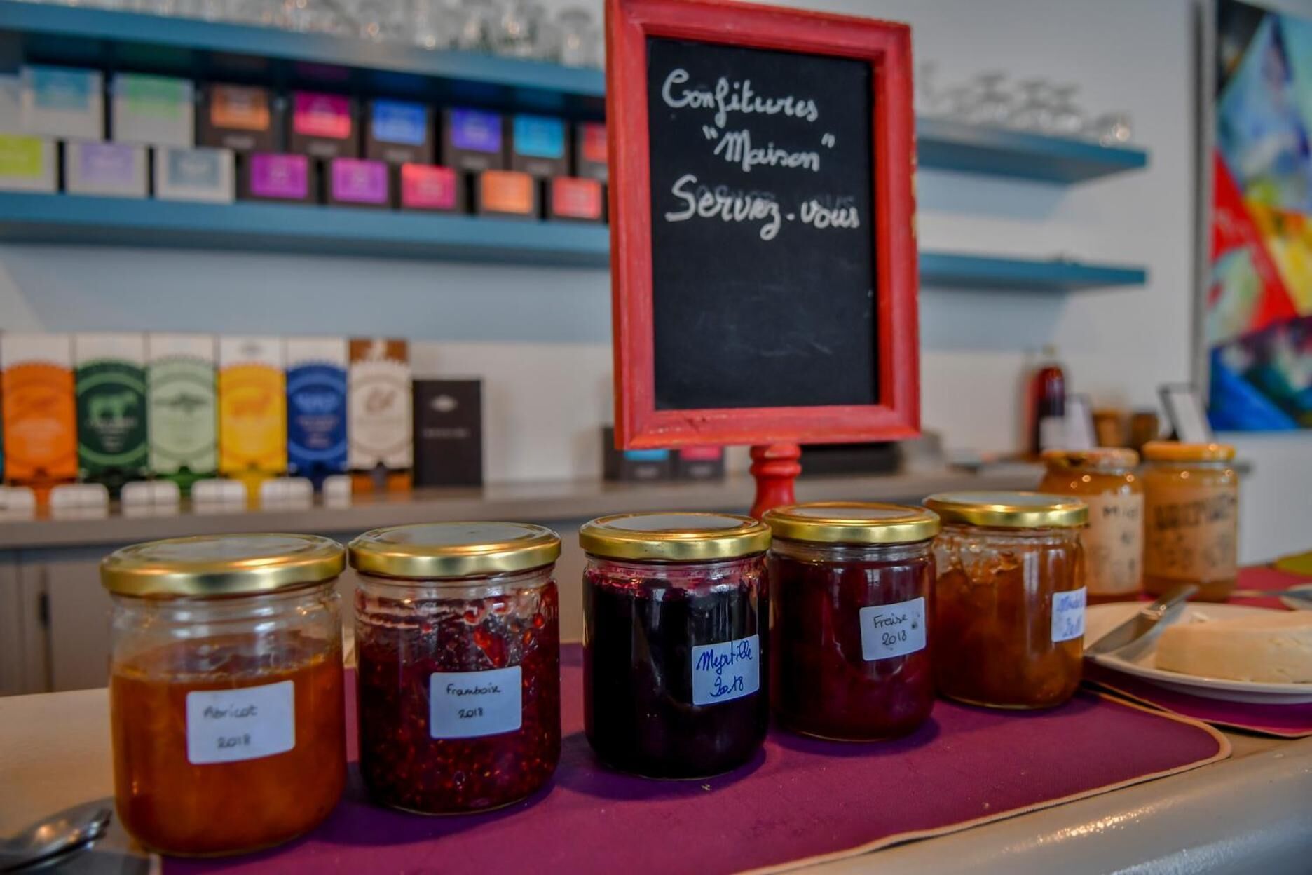On peut voir une variété de pots de confitures et de conserves, chacun avec son étiquette, un panneau noir et, au fond, des étagères remplies de produits emballés.