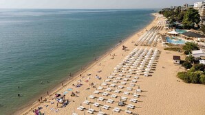 A beach with rows of sun loungers and umbrellas, a pool area, and buildings in the background. - Palm Beach Hotel- All Inclusive (Golden Sands)