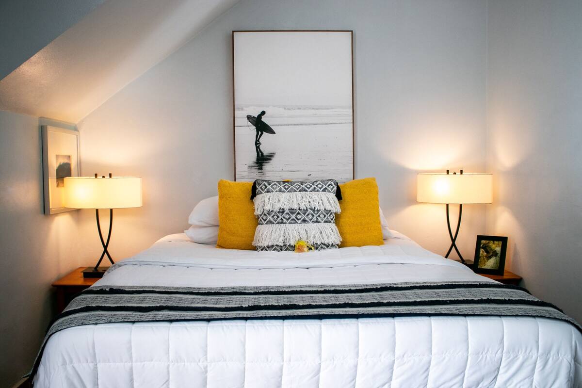Cama bem arrumada com um edredom branco, duas almofadas amarelas e um cobertor com padrão preto e branco. Duas luminárias de cabeceira em cada lado da cama. Um quadro emoldurado na parede acima da cama.